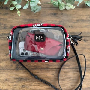 May include: A clear crossbody bag with a red and black plaid trim. Inside, a red wallet with the initials "MS" and a smartphone are visible. The bag has a black strap and a black tassel. The bag is on a wooden surface.