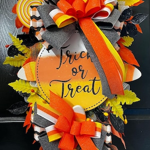 May include: A Halloween wreath with a candy corn theme. The wreath features a round sign that says "Trick or Treat" in black script. It is decorated with orange, yellow, white, and black ribbons, candy corn accents, and decorative leaves.