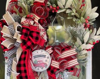 May include: A Christmas wreath with red and black buffalo plaid ribbon, red and white striped ribbon, and a burlap bow. The wreath is decorated with greenery, berries, and a metal sign that says "Farm Fresh Christmas Trees" with a red truck and a Christmas tree.