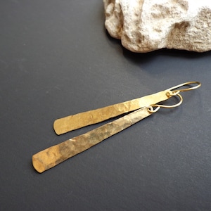 Peut inclure: Une paire de boucles d'oreilles en métal martelé de couleur or, de forme rectangulaire allongée. Les boucles d'oreilles sont suspendues à une fine boucle de fil.