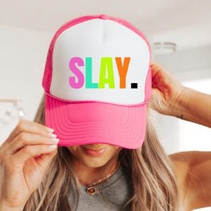 May include: A pink trucker hat with a white front panel. The hat features the word "SLAY" in colorful letters. The hat has a mesh back and a curved brim.