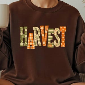 以下が含まれることがあります： 胸元に「HARVEST」の文字がプリントされたダークブラウンのクルーネックトレーナー。各文字には、緑の花柄、オレンジのギンガムチェック、オレンジの花柄、カボチャが描かれた緑のストライプ、オレンジの水玉模様など、それぞれ異なるパターンが施されています。