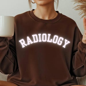 Op de afbeelding: Een bruine sweatshirt met het woord "RADIOLOGY" in witte, gebogen letters. De sweatshirt is effen van kleur en heeft lange mouwen. Een witte mok wordt in de linkerhand gehouden.