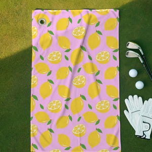 May include: A rectangular golf towel featuring a repeating pattern of yellow lemons and lemon slices on a light pink background. The towel includes a silver metal grommet in the top left corner for attachment to a golf bag.