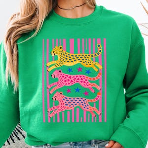 Op de afbeelding: Groene sweatshirt met een afbeelding van drie rennende cheeta's in geel, roze en blauw, tegen een roze gestreepte achtergrond. Blauwe sterren zijn verspreid tussen de cheeta's. De sweatshirt is effen groen.