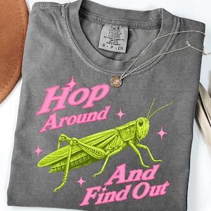 May include: A charcoal gray short-sleeve t-shirt featuring a bright green grasshopper illustration centered on the chest. The text "Hop Around And Find Out" is printed in a bold pink font, accented by small pink four-pointed stars surrounding the insect.