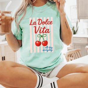 Puede incluir: Camiseta verde claro con las palabras "La Dolce Vita" en rojo, un par de cerezas rojas y la frase "all day everyday" en azul. La camiseta tiene rayas verticales rosas y blancas.