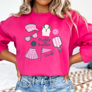 Women’s crewneck sweatshirt featuring the phrase “Pickle Baller” in bold lettering, decorated with playful pickleball-themed graphics such as paddles, balls, and court icons. The design has a sporty, fun vibe perfect for pickleball enthusiasts