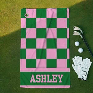 Puede incluir: Una toalla de golf con un diseño de tablero de ajedrez rosa y verde. La toalla tiene el nombre "ASHLEY" impreso en letras rosas grandes en la parte inferior. Palos de golf, pelotas de golf y un par de guantes de golf blancos.