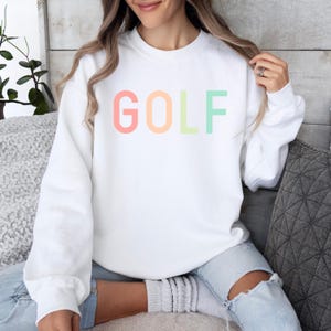 Pode incluir: Camisola branca com a palavra "GOLF" impressa em cores do arco-&iacute;ris.