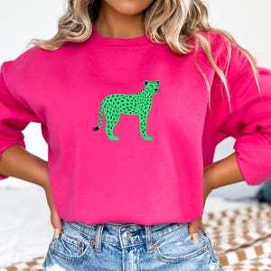 Puede incluir: Una sudadera rosa brillante con un diseño de guepardo verde. El guepardo tiene manchas negras y está de pie. La sudadera tiene mangas largas y cuello redondo. El diseño está centrado en la parte delantera.