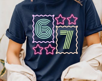 May include: Navy blue t-shirt featuring the numbers "6" and "7" in a retro neon style. The number "6" is in a teal and green color, and the number "7" is in a yellow and green color. Pink stars surround the numbers.