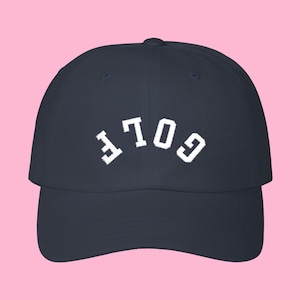 Puede incluir: Gorra de béisbol azul marino con la palabra blanca "GOLF" arqueada en la parte delantera. La gorra tiene una visera curva y una corona redondeada. El fondo es de color rosa liso.