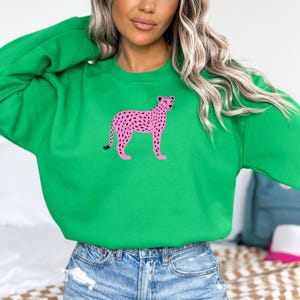 Puede incluir: Una sudadera verde brillante con un diseño de guepardo rosa con manchas negras. El guepardo está de pie con las patas separadas. La sudadera está hecha de un material suave y es adecuada para uso casual.