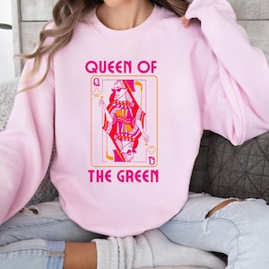 May include: A light pink sweatshirt featuring the text "QUEEN OF THE GREEN" above a playing card design. The card depicts a woman in sunglasses with a red and pink outfit. The design is outlined in orange.