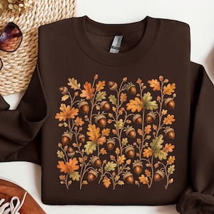 Op de afbeelding: Een donkerbruine sweater met ronde hals en een centrale grafische print van herfstachtige eikenbladeren en eikels in tinten oranje, geel en groen. Het kledingstuk met lange mouwen heeft een geribbelde kraag, boorden en een zoom.