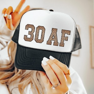May include: A black and white trucker hat with the text "30 AF" in a leopard print design. The hat has a mesh back and a curved brim. The hat is a fun accessory for a 30th birthday celebration.