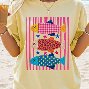 Puede incluir: Una camiseta de color amarillo pálido con un estampado gráfico de tres peces estilizados apilados verticalmente. El pez superior tiene un patrón de cuadros rosa, el del medio un estampado de leopardo naranja y el inferior es azul con lunares amarillos.