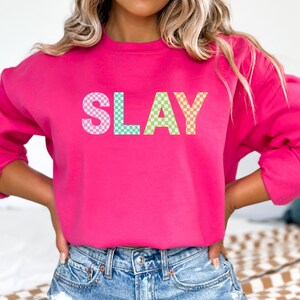 May include: A bright pink sweatshirt with the word "SLAY" in large, checkered letters across the front. The letters are in pink, blue, green, and orange. The sweatshirt has long sleeves and a crew neck.
