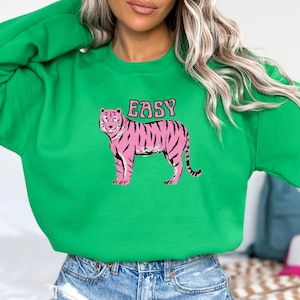 Puede incluir: Sudadera verde con una ilustración de tigre rosa y la palabra "EASY". El tigre tiene rayas negras y un collar. El texto está en una fuente retro. La sudadera es de color verde liso.