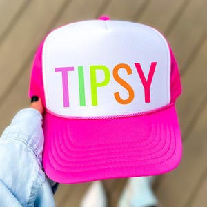 May include: A bright pink trucker hat with a white front panel. The word "TIPSY" is printed across the front in colorful letters. The hat has a mesh back and a curved brim. A fun and stylish accessory.