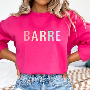 May include: A bright pink sweatshirt with the word "BARRE" printed across the chest in pastel rainbow colors. The sweatshirt has long sleeves and a crew neck. The model is wearing blue jeans.