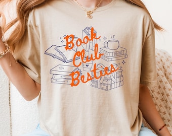 May include: Beige t-shirt featuring a graphic design with the text "Book Club Besties" in orange script. The design includes illustrations of books, a coffee cup, and a stack of gifts, all outlined in blue.