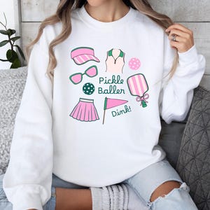 May include: White sweatshirt with a pink and green pickleball design. The design includes a pickleball paddle, a ball, a visor, a skirt, a shirt, sunglasses, and a flag with the word "Dink!" on it. The text "Pickle Baller" is also included in the design.