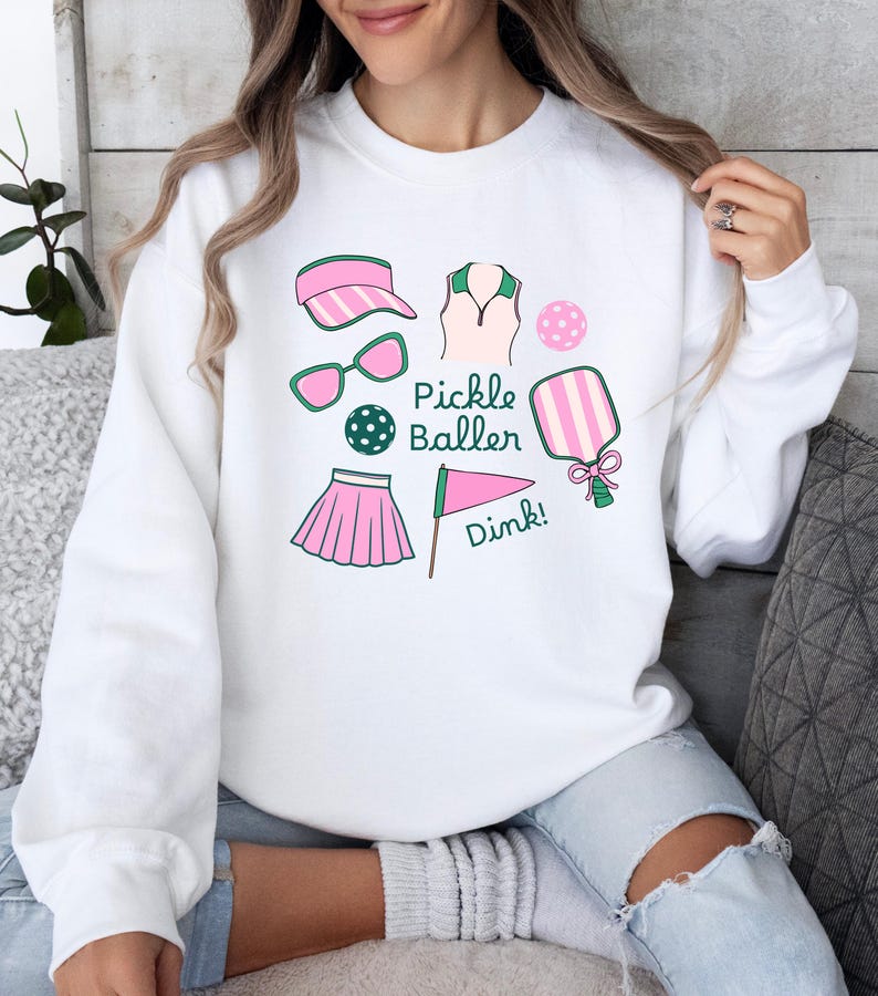 May include: White sweatshirt with a pink and green pickleball design. The design includes a pickleball paddle, a ball, a visor, a skirt, a shirt, sunglasses, and a flag with the word "Dink!" on it. The text "Pickle Baller" is also included in the design.