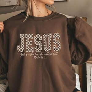 Può includere: Una felpa marrone con la parola "JESUS" in un motivo a scacchi sul petto. Sotto, il testo "God is within her; she will not fall Psalm 46:5".