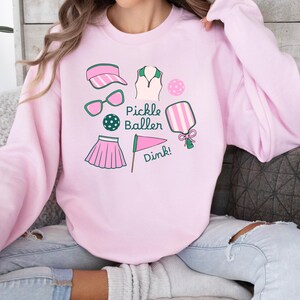 May include: Pink sweatshirt with a graphic design of pickleball equipment, including a paddle, ball, visor, sunglasses, skirt, and a flag with the text "Pickle Baller Dink!"