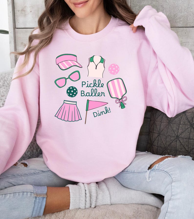 May include: Pink sweatshirt with a graphic design of pickleball equipment, including a paddle, ball, visor, sunglasses, skirt, and a flag with the text "Pickle Baller Dink!"