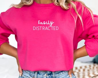 Może przedstawiać: Różowa bluza z napisem "Easily Distracted" w białym kolorze.