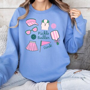 May include: A light blue sweatshirt with a graphic design of pickleball equipment. The design includes a paddle, a ball, sunglasses, a visor, a shirt, a skirt, and a flag. The text "Pickle Baller Dink!" is included in the design.