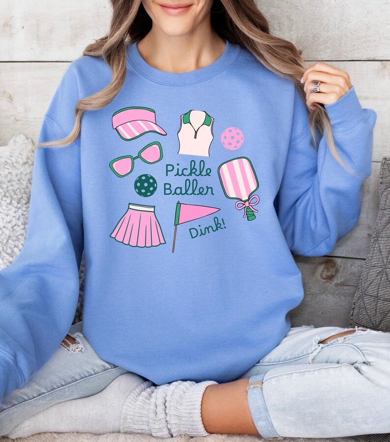 May include: A light blue sweatshirt with a graphic design of pickleball equipment. The design includes a paddle, a ball, sunglasses, a visor, a shirt, a skirt, and a flag. The text "Pickle Baller Dink!" is included in the design.