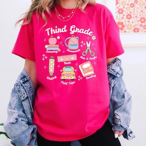 Puede incluir: Una camiseta rosa intenso de Tercer Grado con gráficos coloridos con temática escolar. El diseño incluye una manzana, una mochila, tijeras, libros, un lápiz, una regla y un autobús escolar. Las palabras "Third Grade" están en una fuente caprichosa.