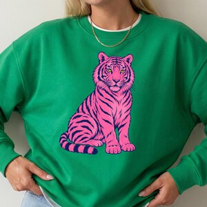Puede incluir: Una sudadera de cuello redondo en color verde vibrante con una ilustración gráfica de un tigre sentado. El tigre está representado en rosa brillante con rayas moradas oscuras y ojos amarillos. La sudadera tiene mangas largas y un cuello acanalado.