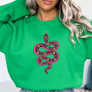 Puede incluir: Una sudadera de cuello redondo en color verde brillante con un gráfico central de una serpiente enroscada. La serpiente es de color rosa vibrante con un patrón interior negro lleno de pequeñas flores amarillas y detalles de líneas negras.