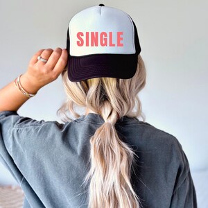 Puede incluir: Una gorra de camionero negra y blanca con un gráfico rojo "SINGLE" en la parte delantera. La gorra la lleva una persona con el pelo largo y rubio.