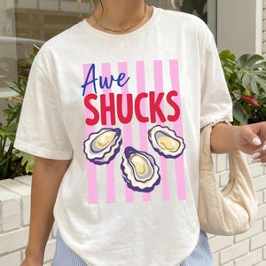 Puede incluir: Camiseta blanca con el texto "Awe Shucks" en azul y rojo, con tres ilustraciones de ostras. Rayas verticales rosas detrás del texto y las ostras. La camiseta es de estilo casual y cómodo.