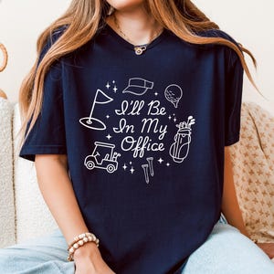 Op de afbeelding: Marineblauw t-shirt met een wit grafisch ontwerp met golfgerelateerde iconen en de tekst "I'll Be In My Office". Het ontwerp bevat een golflag, vizier, golfbal, golftas, golfkar en tees.
