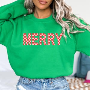May include: A bright green sweatshirt with the word "MERRY" in a red and white checkered pattern across the chest. The sweatshirt is a solid color and has long sleeves. The model is wearing blue jeans.