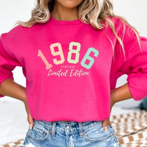 May include: A bright pink sweatshirt with the year "1986" in pastel colors and the words "Vintage Limited Edition". The sweatshirt is a crew neck style.