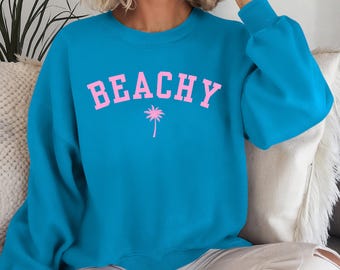 Può includere: Una felpa turchese con la scritta "BEACHY" in lettere rosa e una grafica di palma rosa. La felpa ha maniche lunghe e girocollo.