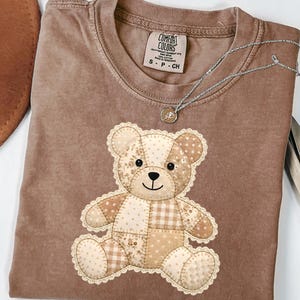May include: A brown Comfort Colors t-shirt featuring a patchwork teddy bear design. The bear is made of various beige and tan fabrics with a lace trim. A silver necklace with a small pendant is draped over the shirt.