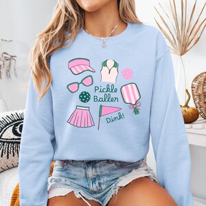 May include: Light blue sweatshirt with a graphic design of pickleball equipment, including a paddle, ball, visor, sunglasses, skirt, and a flag with the text "Pickle Baller Dink!"