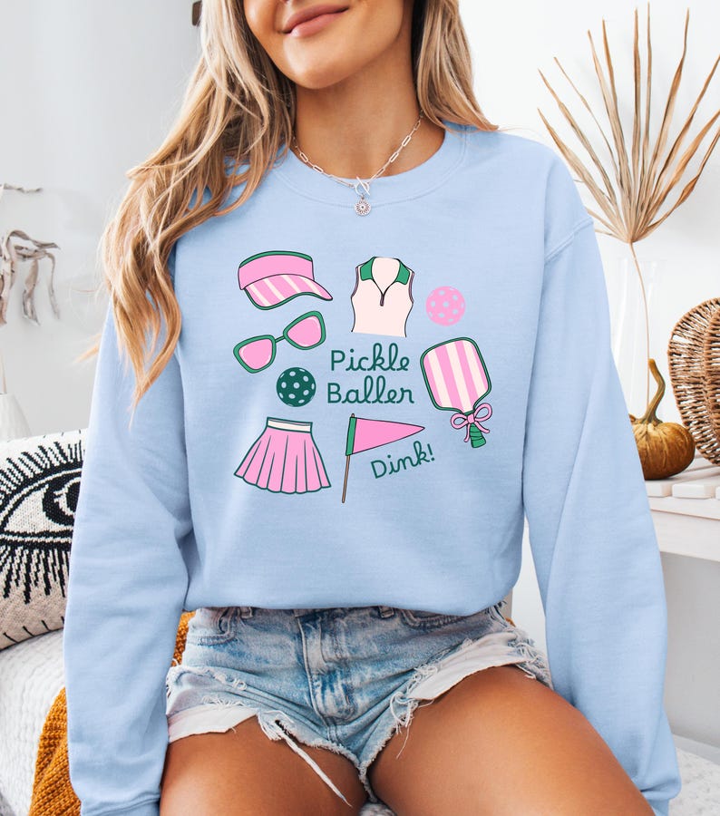 May include: Light blue sweatshirt with a graphic design of pickleball equipment, including a paddle, ball, visor, sunglasses, skirt, and a flag with the text "Pickle Baller Dink!"