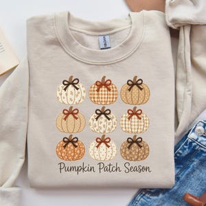 May include: Beige sweatshirt featuring a grid of nine decorative pumpkins in various patterns and colors, each with a bow. The text "Pumpkin Patch Season" is printed below the pumpkins. The sweatshirt is folded on a white surface.