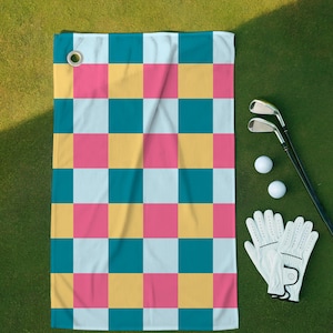 Puede incluir: Una toalla de golf con un patrón de tablero de ajedrez colorido en turquesa, rosa, amarillo y blanco. Dos pelotas de golf, un par de palos de golf y un par de guantes de golf blancos están junto a la toalla sobre una superficie verde.