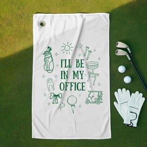 Op de afbeelding: Witte golfhanddoek met groene illustraties van golfartikelen en de tekst "I'll be in my office". Het ontwerp omvat een golftas, een golfkar en een golfbal. Golfclubs, ballen en handschoenen zijn ook zichtbaar.